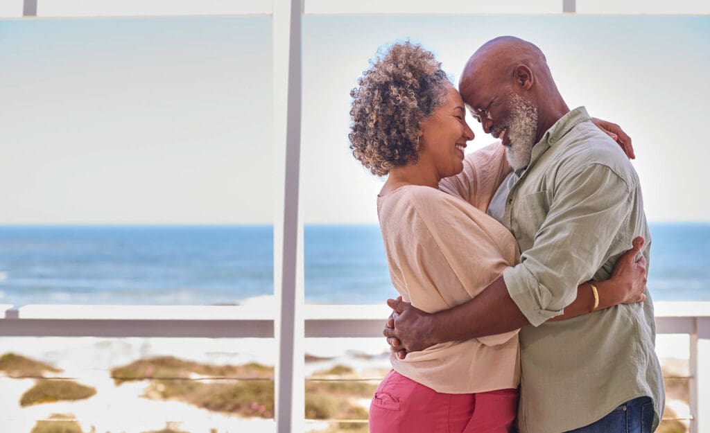 Loving Mature Couple Hugging In Beachfront House Overlooking Ocean For Summer Vacation