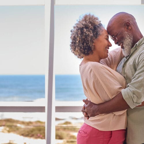 Loving Mature Couple Hugging In Beachfront House Overlooking Ocean For Summer Vacation