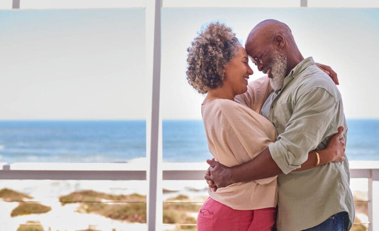 Loving Mature Couple Hugging In Beachfront House Overlooking Ocean For Summer Vacation
