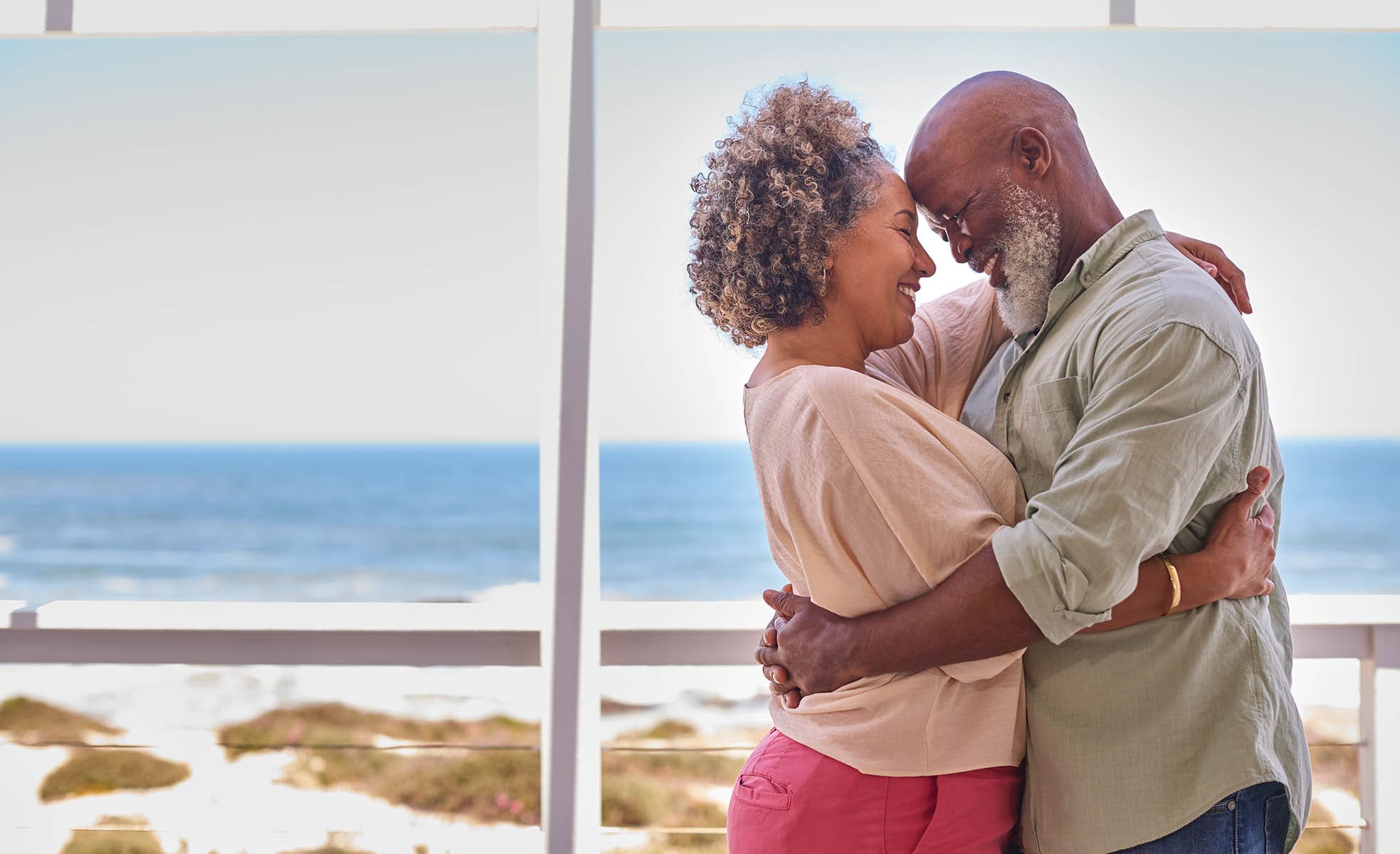 Loving Mature Couple Hugging In Beachfront House Overlooking Ocean For Summer Vacation