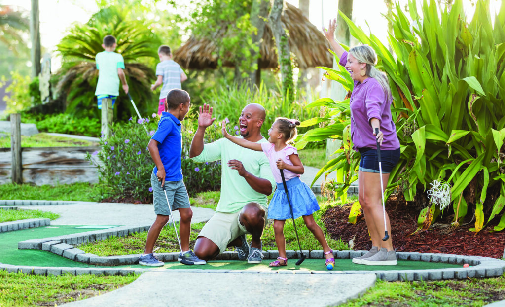 An interracial family having fun together playing miniature golf. The African-American father and Caucasian mother are in their 30s. The children are 9 and 5 years old. Dad and sister are giving the boy high fives for his winning putt.