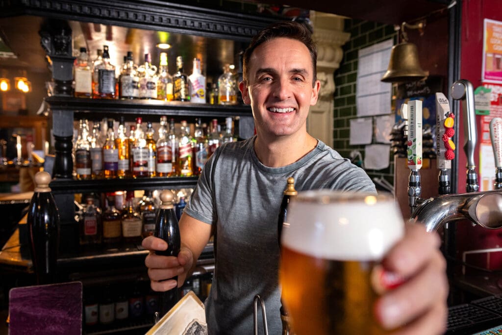 Waist-up shot of a mid-adult male bar worker looking into the camera, holding a beer at the camera. He is behind the bar surrounded by numerous lagers and beers. The bar/restaurant is located in Hexham.